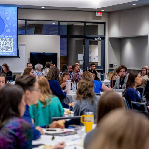at GVSU CommCon, people seated at round tables, screens show an agenda with Fireside Chat: President Mantella and Vice President Stacie Behler.